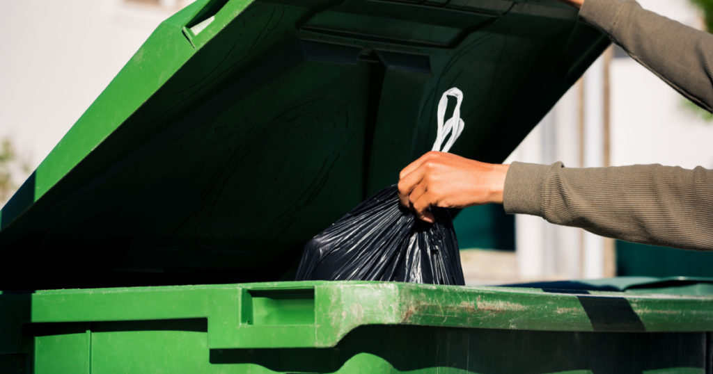 Man throwing out black eco-friendly recyclable trash bag in to big plastic green garbage container. Take out the trash
