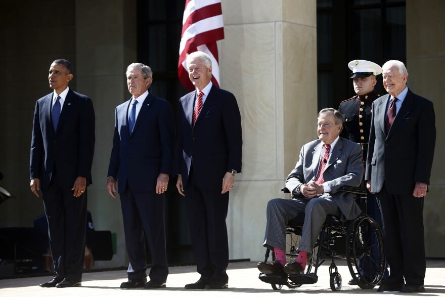 Five former presidents (Barack Obama, George W. Bush, Bill Clinton, George H.W. Bush and Jimmy Carter) are seen standing next to each other with a Marine behind them.