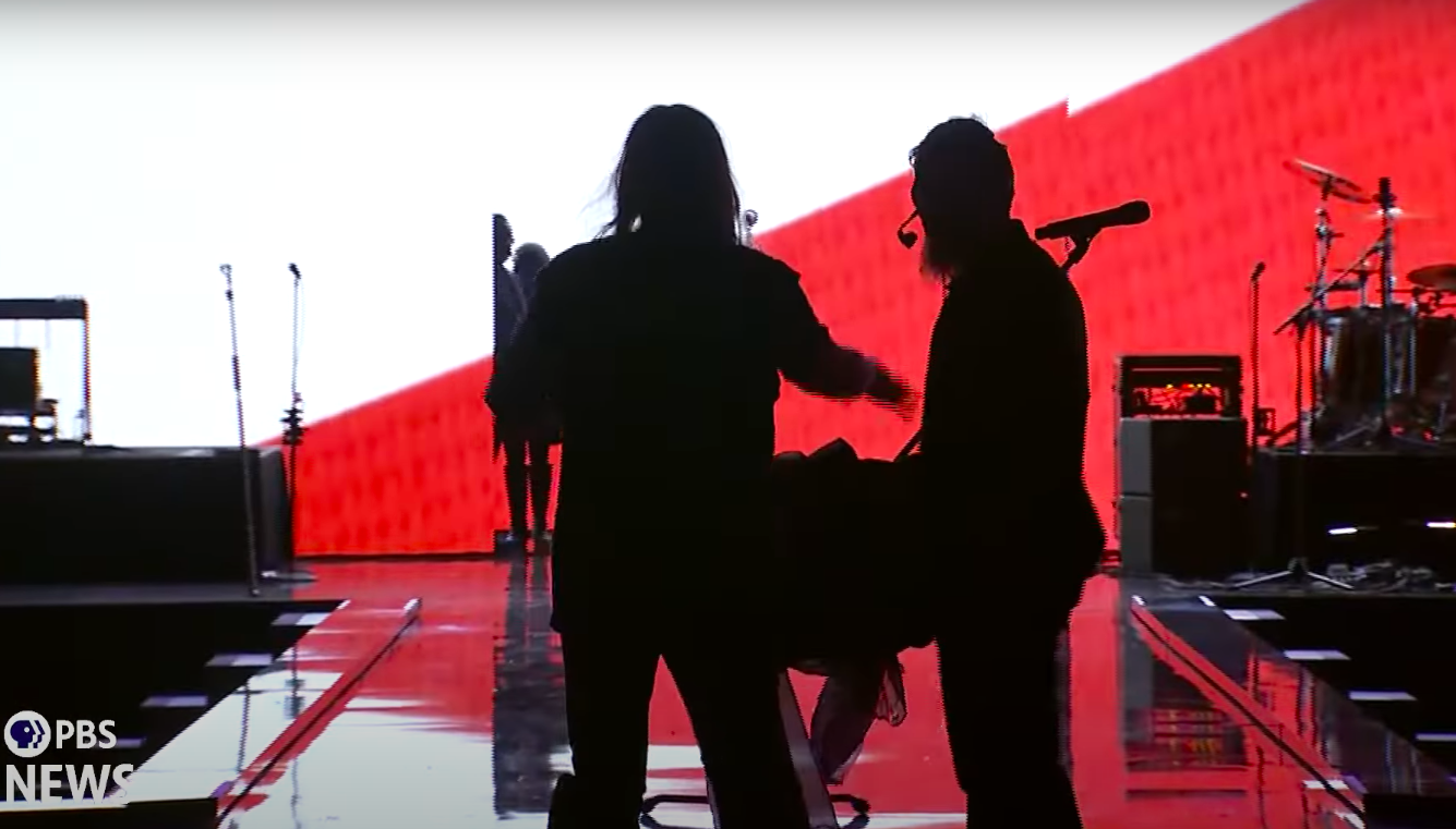 A stagehand helping Billy Ray Cyrus gather his things and exit the stage. | Source: YouTube/PBS NewsHour