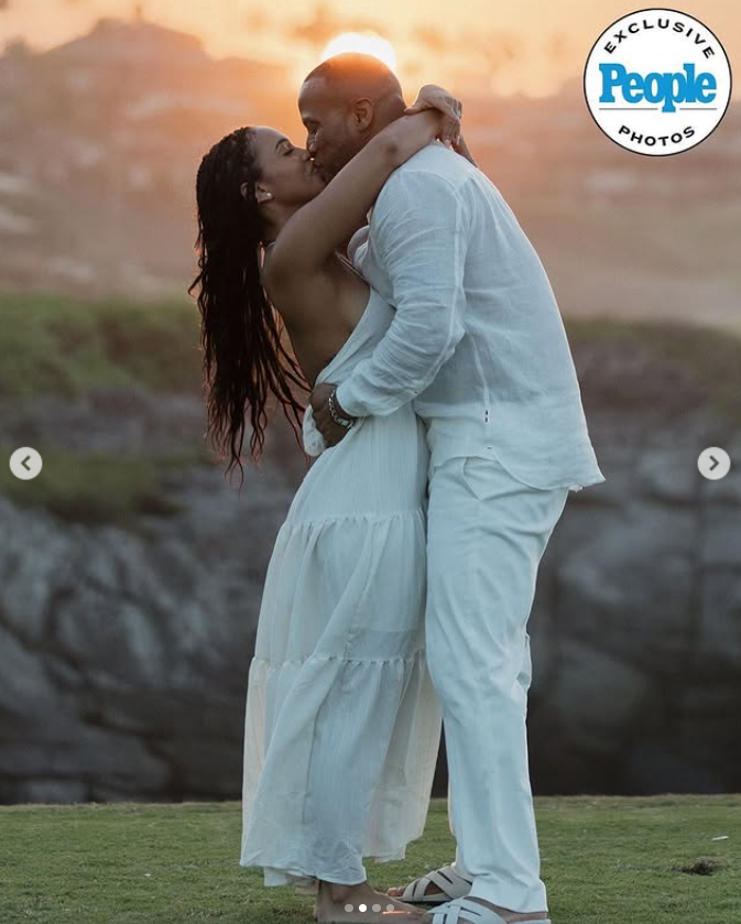 DeVon Franklin and Maria Castillo share a romantic kiss at sunset, embracing on a grassy cliff while dressed in elegant white outfits | Source: Instagram/people