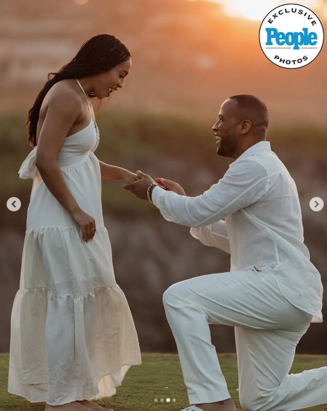 Devon Franklin kneels on one knee, holding Maria Castillo's hand as she smiles in surprise during his heartfelt proposal at sunset | Source: Instagram/people